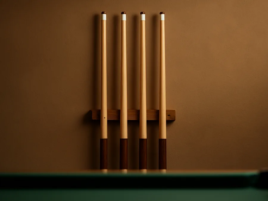 Four maple pool cues standing in a wall rack above a pool table